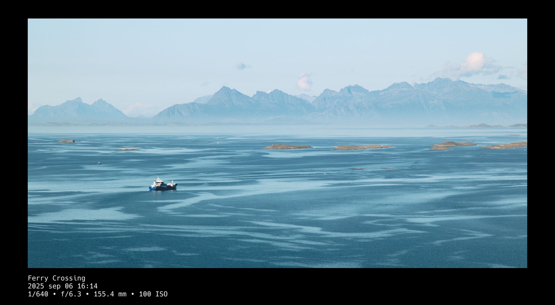 A ferry sailing toward the horizon in a calm, turquoise-blue ocean. Mountains in the far distance are towering over the scene. A black frame surrounds the photo whose title and camera setting details are written in white text beneath it.