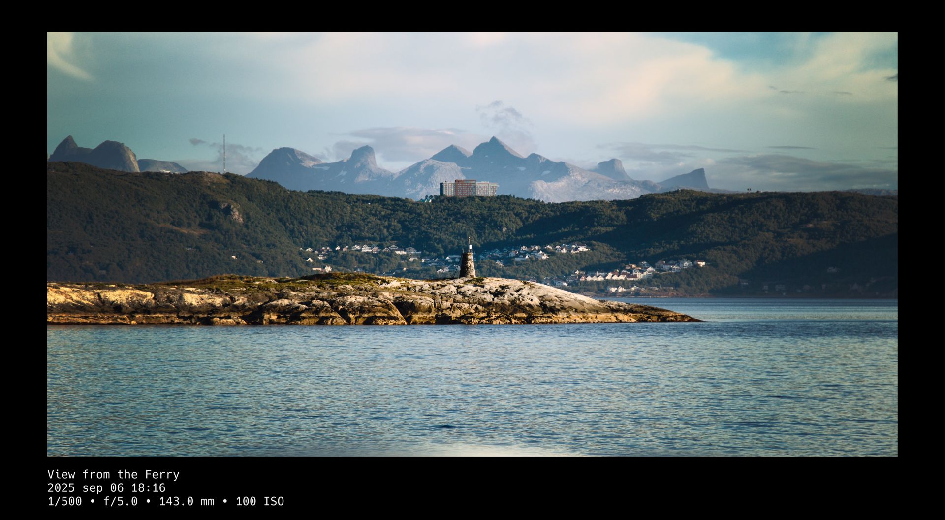 View over ocean water past a lantern on a rocky island, toward a small town resting in the tree covered slope of a large hill. A multi-story building stands atop the hill and a cold, rocky mountain fills the gap toward the background of whispy clouds in a blue sky. A black frame surrounds the photo whose title and camera setting details are written in white text beneath it.