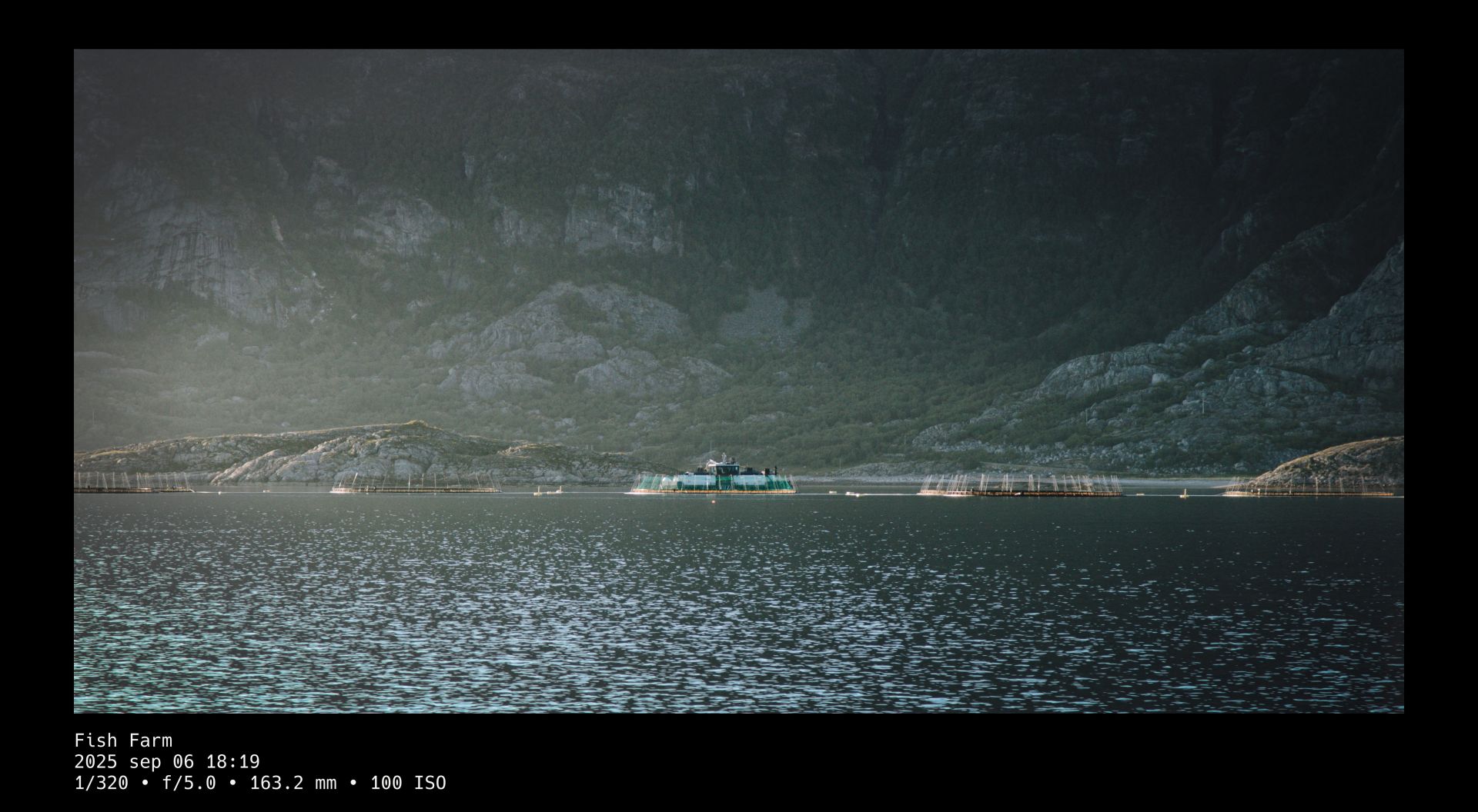 View over ocean water toward a fisherboat tending to several circular fish-farm pods in front of a massive, rocky mountain side overgrown with bushes and trees. A black frame surrounds the photo whose title and camera setting details are written in white text beneath it.