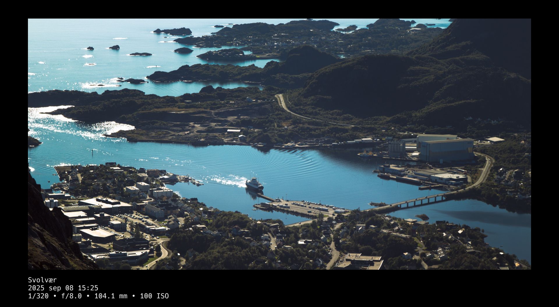 Birdseye view overlooking an ocean-side city harbour, where cars are approaching over a winding road to line up for boarding the approaching ferry. A black frame surrounds the photo whose title and camera setting details are written in white text beneath it.