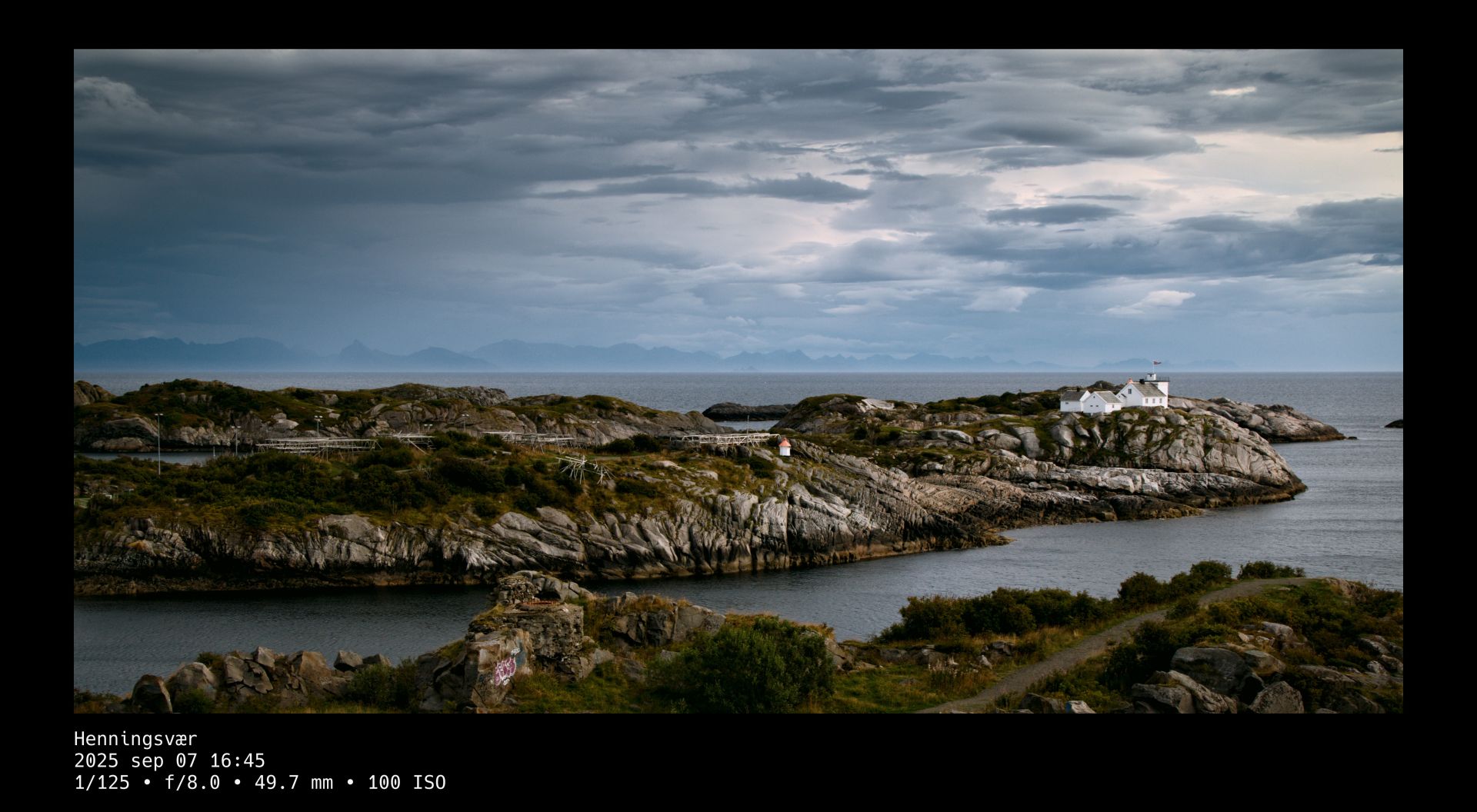 A white house built upon a solid rock foundation by the sea before a dramatic sky. A dark line on the rock shows where the high tide lies when it's in. A black frame surrounds the photo whose title and camera setting details are written in white text beneath it.