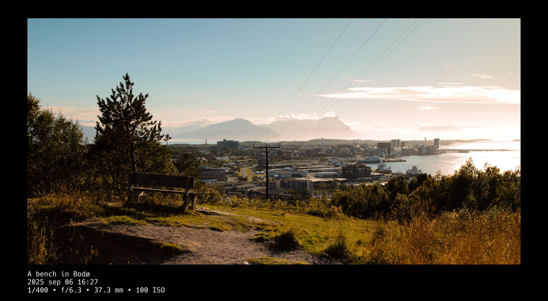 A warm late-summer light is cast upon a wooden bench standing on a grassy hill that overlooks a city. Power lines lead from the top right toward the center of the image and thin, bright clouds and mist softly wrap around the mountains on the horizon. A black frame surrounds the photo whose title and camera setting details are written in white text beneath it.